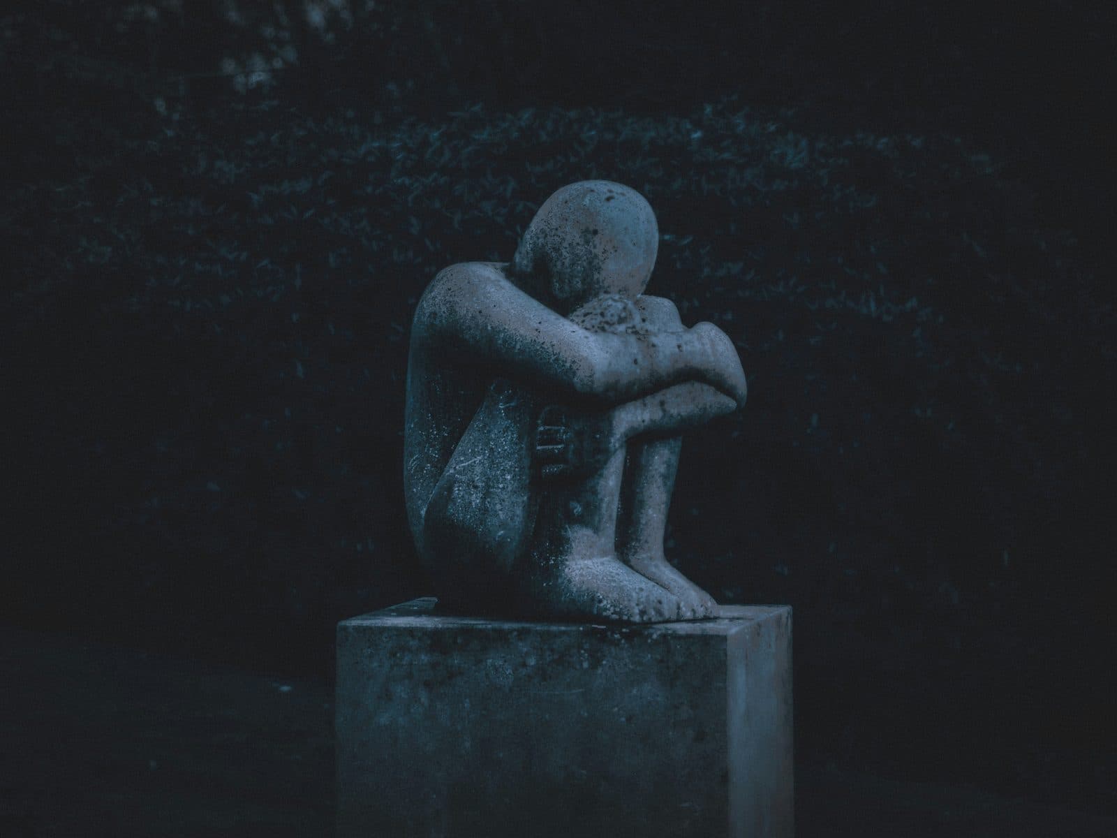 A weathered stone statue of a seated figure curled inward, arms wrapped around its knees