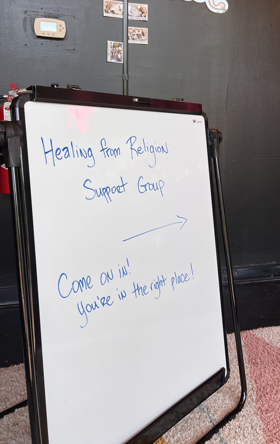 A handwritten whiteboard sign at The Movement Lounge reading “Healing from Religion Support Group — come on in! You’re in the right place!”