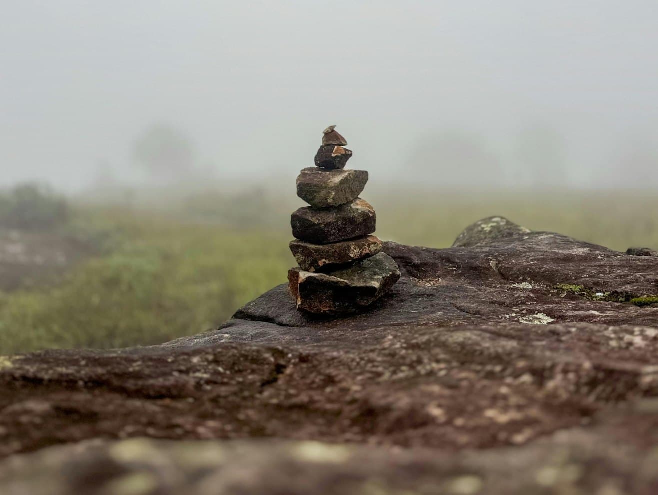 A small cairn of stacked stones on a rock outcrop in morning mist — a trail marker left by someone who came this way before