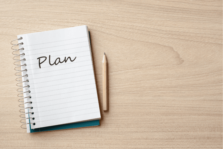 A spiral notebook with the word Plan written on it, next to a pencil on a wooden desk